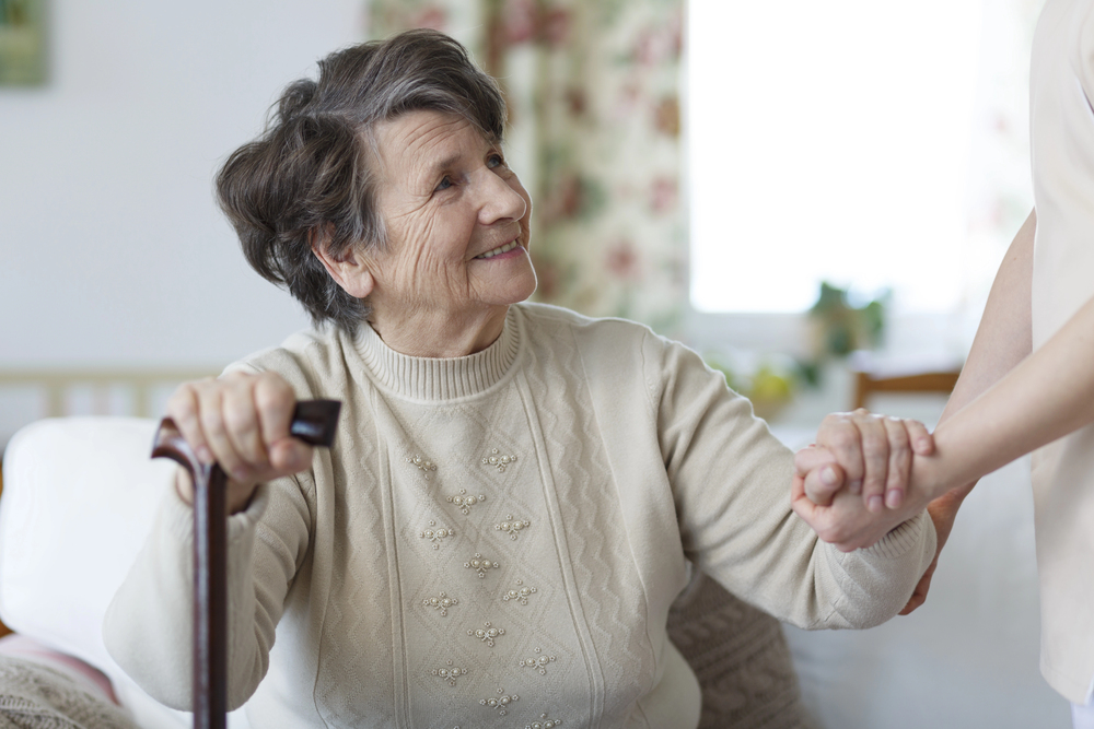 Depositphotos_151099372_S Smiling woman with a cane being helped up by a caregiever.