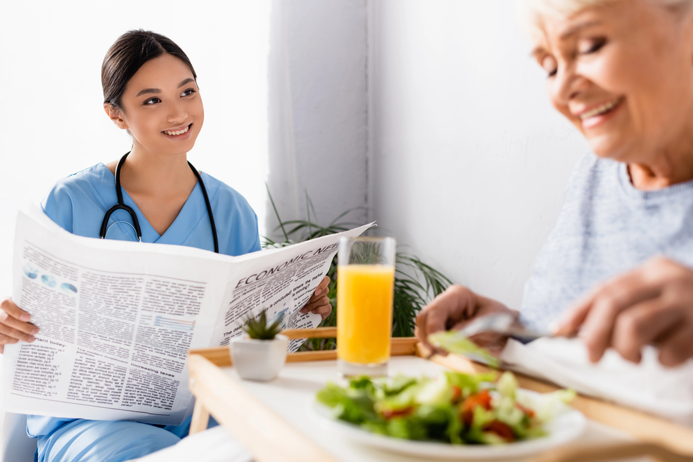 Depositphotos_426798440_S Caregiver reading a newspaper to a senior having breakfast at home